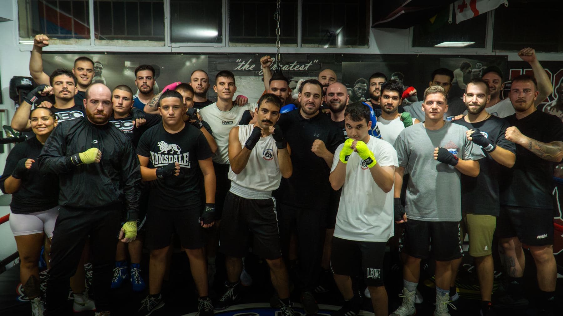 Ολόκληρη η ομάδα του Onomastos Boxing Club στέκεται μαζί στην αίθουσα με τις γροθιές ψηλά, αθλητές κάθε ηλικίας και επιπέδου.